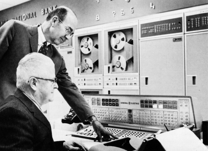 Two men in suits stand and sit in front of large wall of computer equipment. One man points to buttons on console and keyboard.