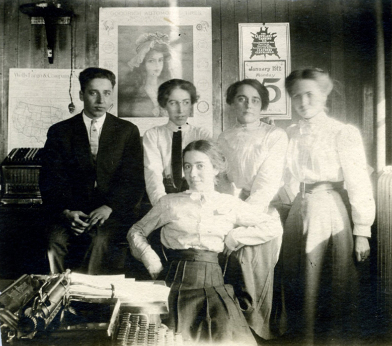 Four women in long skirts and one man sit or stand inside of office room, looking directly at camera. Typewriter in foreground, posters and calendars on wall in background.