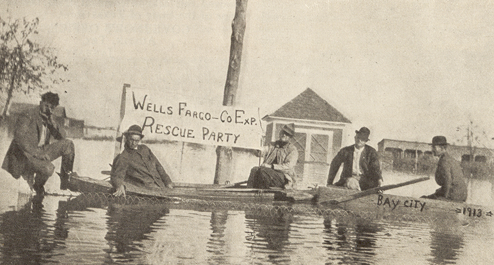 Five men in two rowboats floating in shallow water. Sign says Wells Fargo & Co Exp Rescue Party. Flooded buildings in background. Image is black and white.