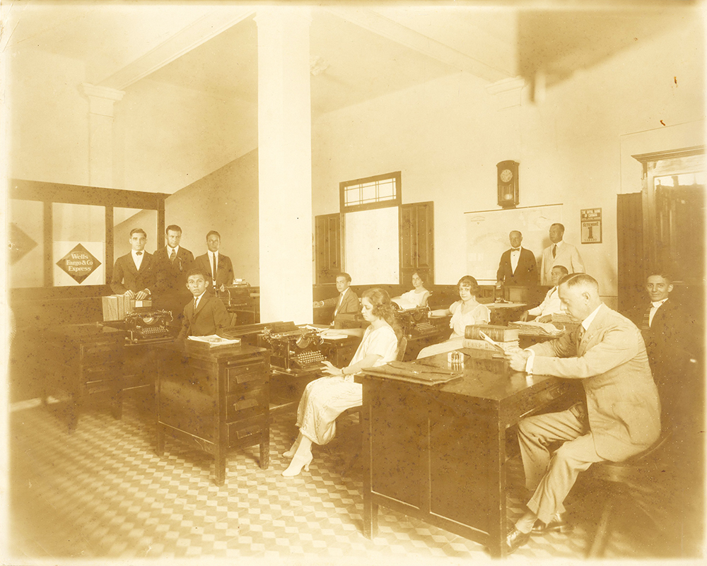 Office interior showing nine men and three women in suits and dresses standing and sitting at desks with typewriters. Wells Fargo & Co Express sign in background.