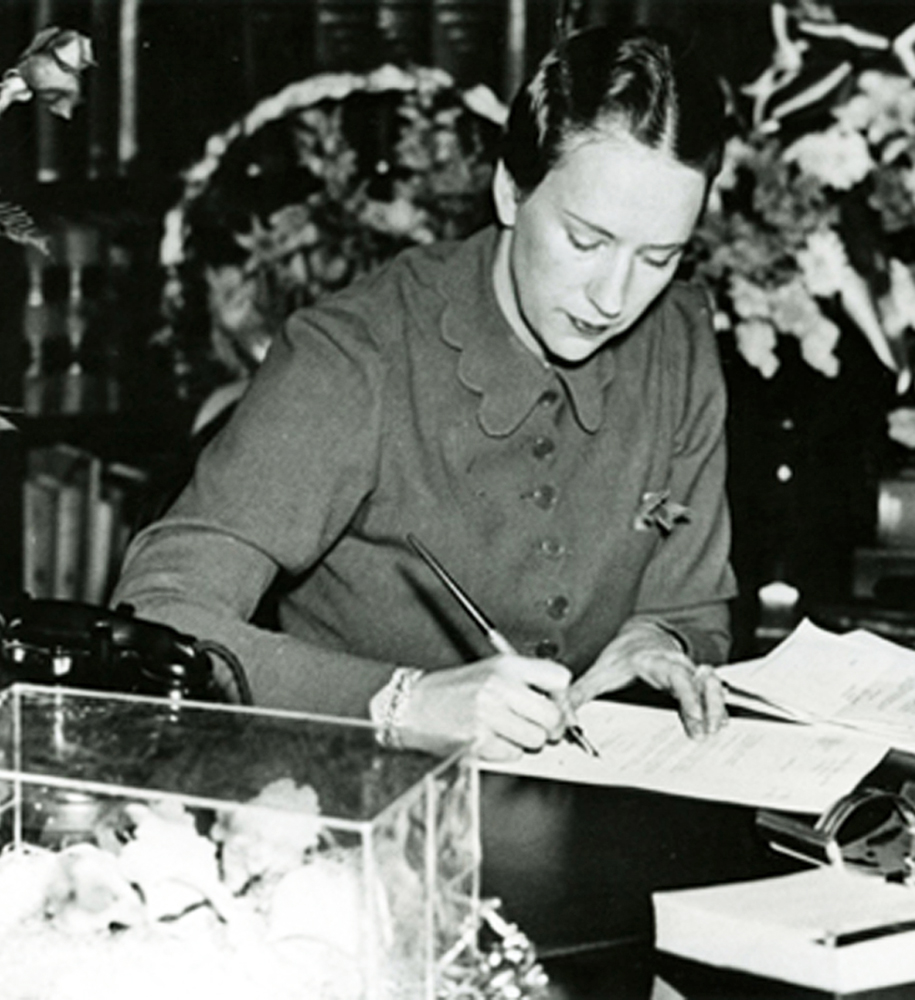 Woman in business attire sits at a large desk signing a stack of papers. Floral displays in background.