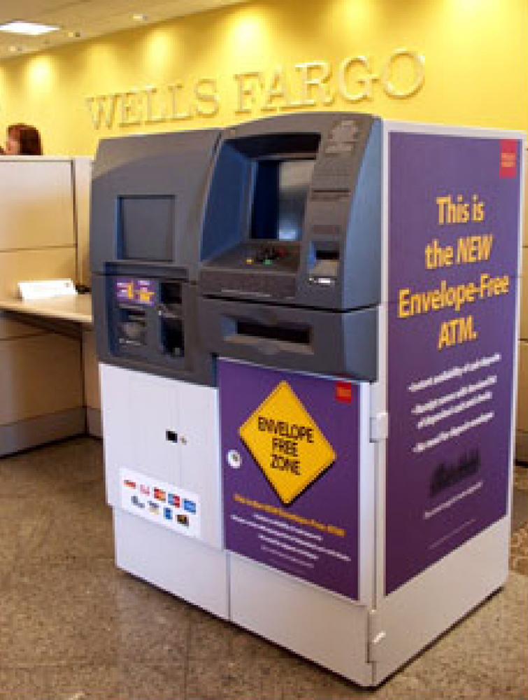 ATM machine in large metal enclosure sits against a wall. Purple signage on side of machine reads this is the new envelope-free ATM. Purple diamond sign on front reads Envelope Free Zone.