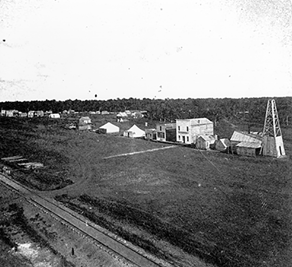 Photo of several wood buildings and canvas tents built on one side of a wide dirt street. Single track railroad track in foreground, trees in background. Photo is black and white.