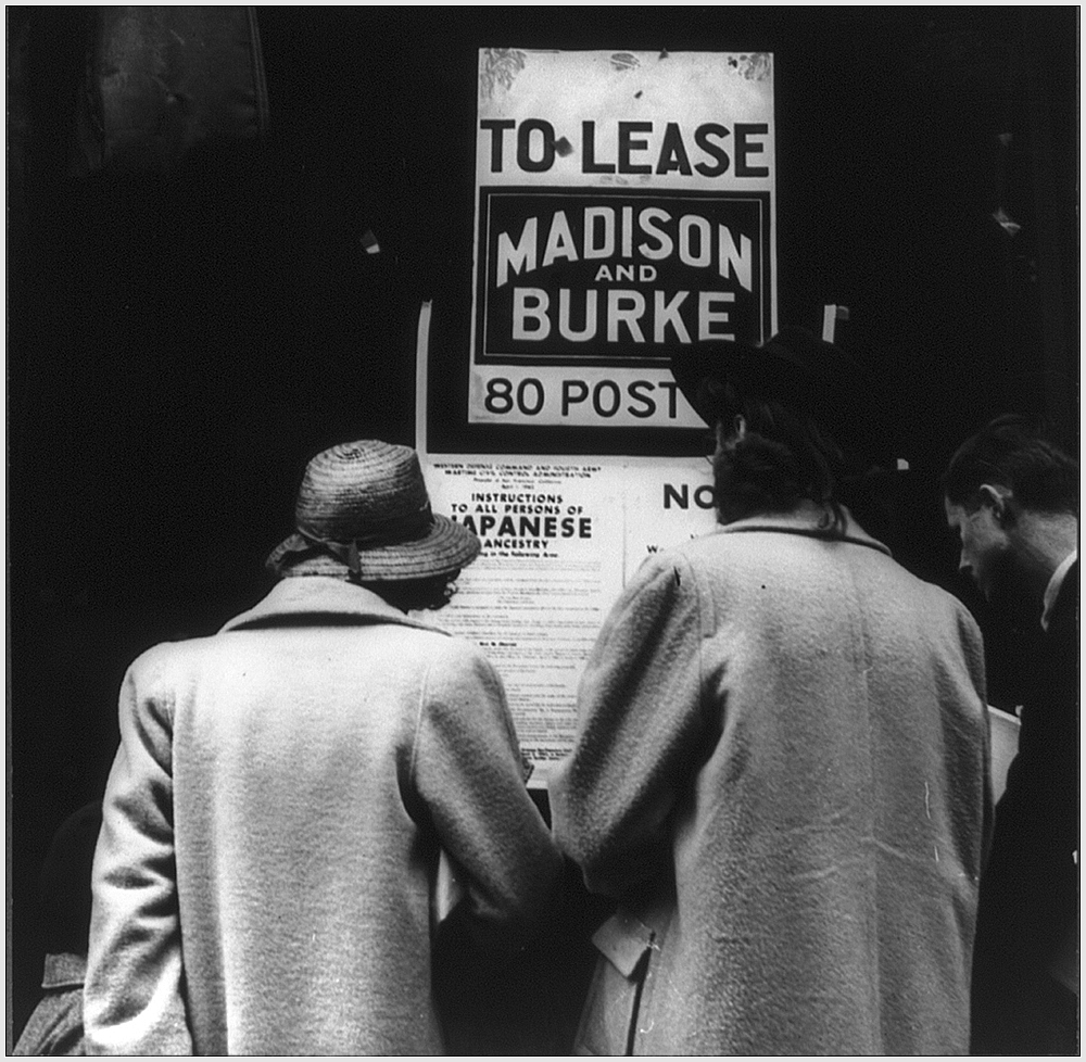 Two women in coats and hats and one man face away from camera reading a notice posted in vacant store window. Notice reads Instructions to all persons of Japanese Ancestry. Additional printing illegible.