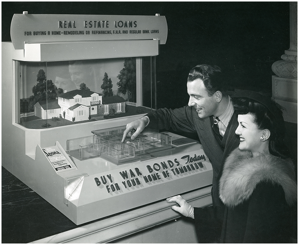 Smiling man and woman stand looking at a display of a scale model of a house and floorplan inside a case. Sign reads Real Estate Loans for buying a home remodeling or refinancing F.H.A. and regular bank loans Buy War Bonds today for your home of tomorrow.