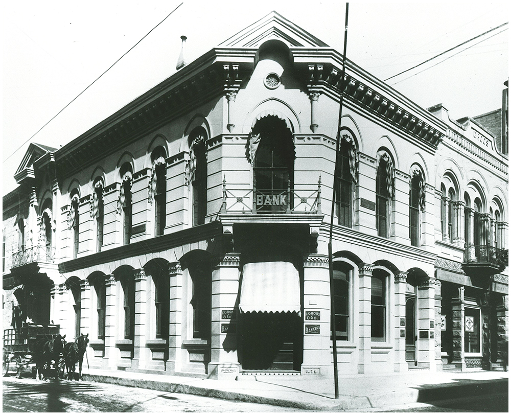 View of two story stone building standing on corner. Over the entrance is sign reading Bank. Photo is taken from street, looking directly at facade.