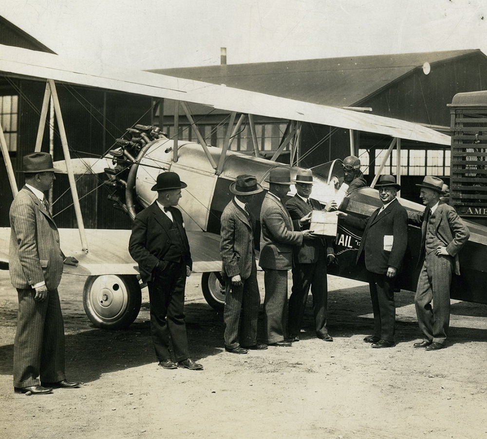 Seven men in suits surrounding a bi-plane on the ground. Two men hand items to the pilot, sitting in an open cockpit. Hangar buildings in background, delivery truck parked at right.