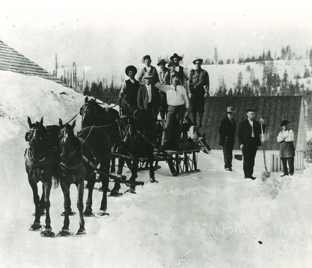 A sled in snow being pulled by four horses wearing flat snowshoes attached to each hoof. Six men and two women stand on sled. Three more men stand to right, one with snow shovel. Buildings in background have snow piled up to roof. Black and white photo.