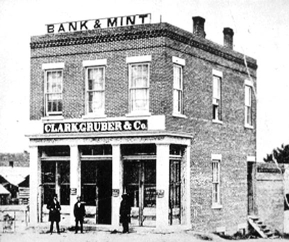 Three men stand outside of small two-story brick building. Sign on rooftop reads bank and mint. Sign over entrance reads Clark, Gruber & Co. Image is black and white.