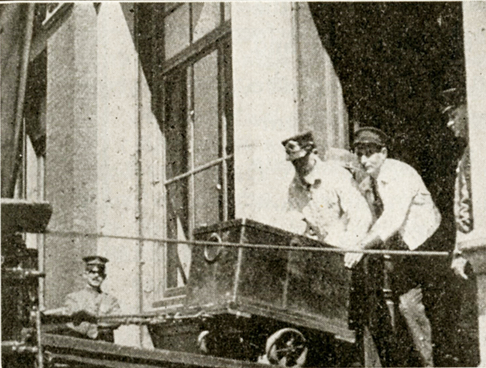 Two men rolling an iron safe from a stone building up a ramp to a waiting wagon. A third man watches from the doorway, and an armed man in military uniform stands guard in background. Black and white photo.
