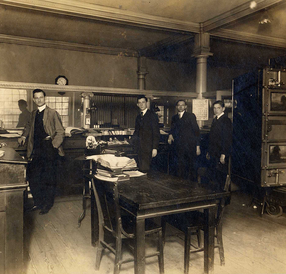 Four men in suits face camera in bank workspace with table, adding machine, documents, and open vault door at right. A calendar on the wall for month of March 1915.