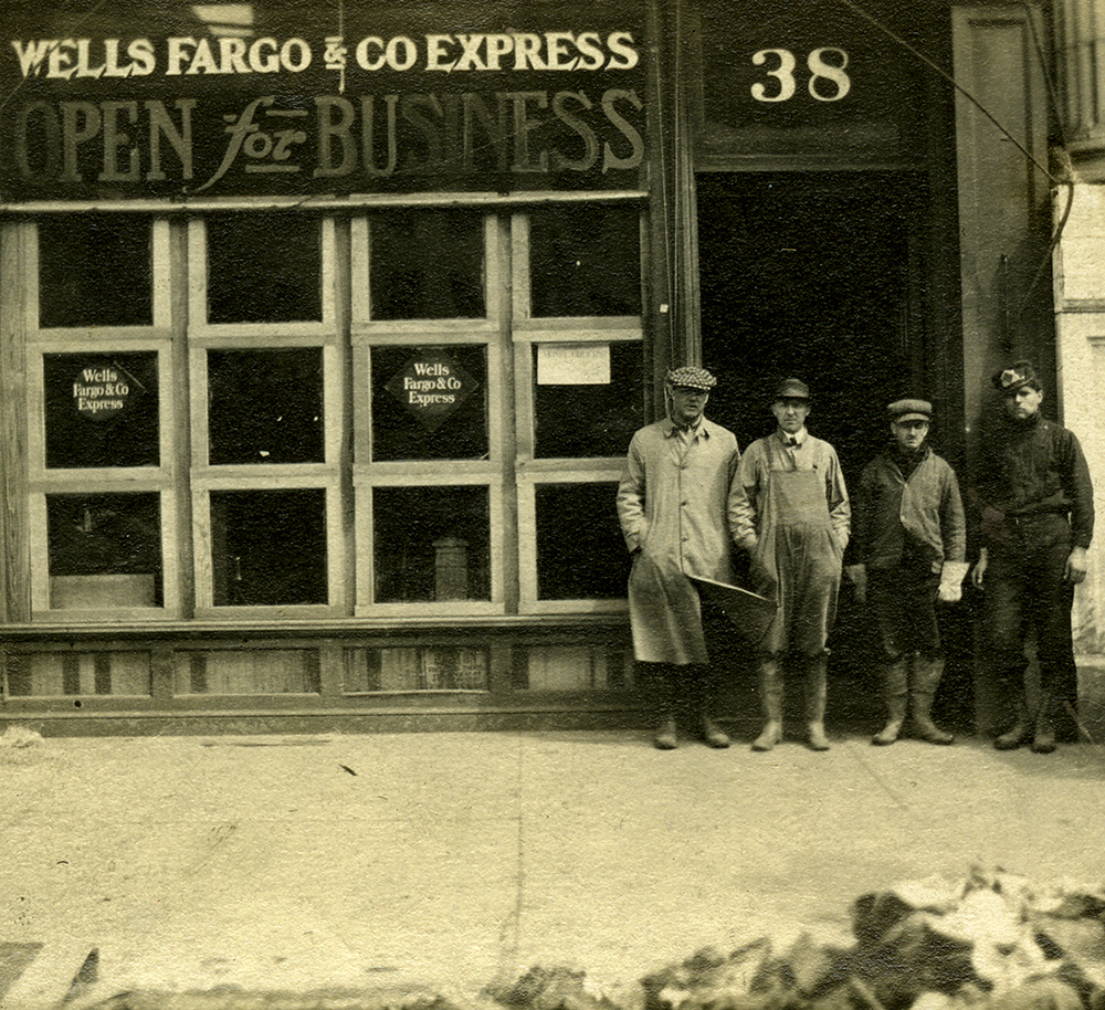 Four men wearing work clothing and rubber boots stand facing the camera on a sidewalk in front of a building. Sign at left says Wells Fargo & Co. Express Open for Business.