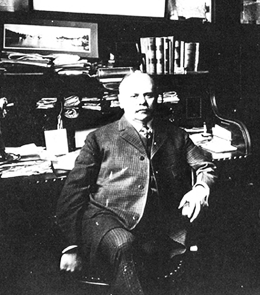 Elderly man in suit sits looking at camera. Behind him is a cluttered roll-top desk full of papers and books. Black and white image.