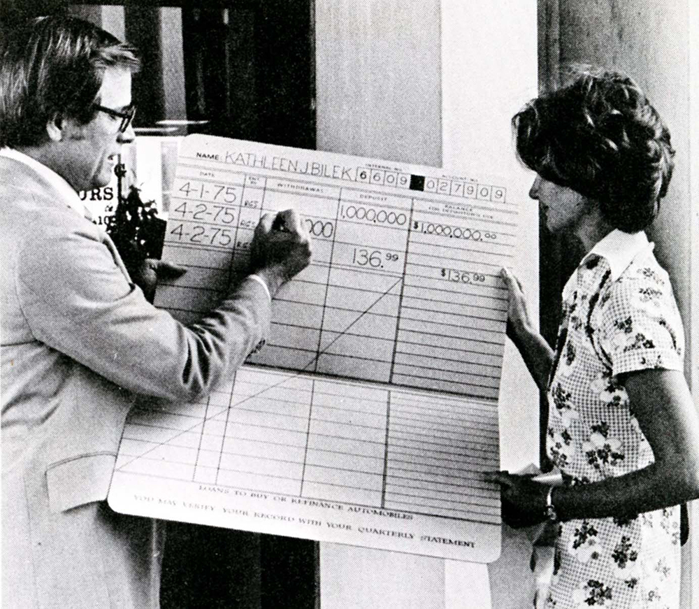Man in suit and woman in dress stand holding oversize savings account passbook between them. Man, at left, writes a number in the passbook.