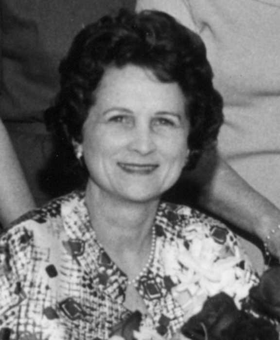 A woman with dark hair looking at camera. In background are other people, unidentified. Black and white image.