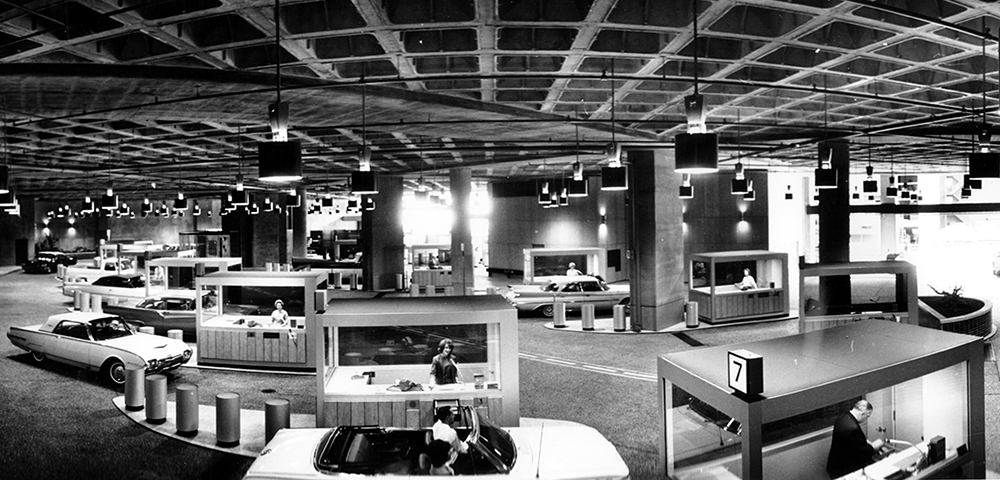 Black and white panoramic photo of drive-in banking facility showing women tellers in multiple metal booths. Cars approach each teller's booth. The roof of the drive-in facility covers the whole scene. Image is black and white.