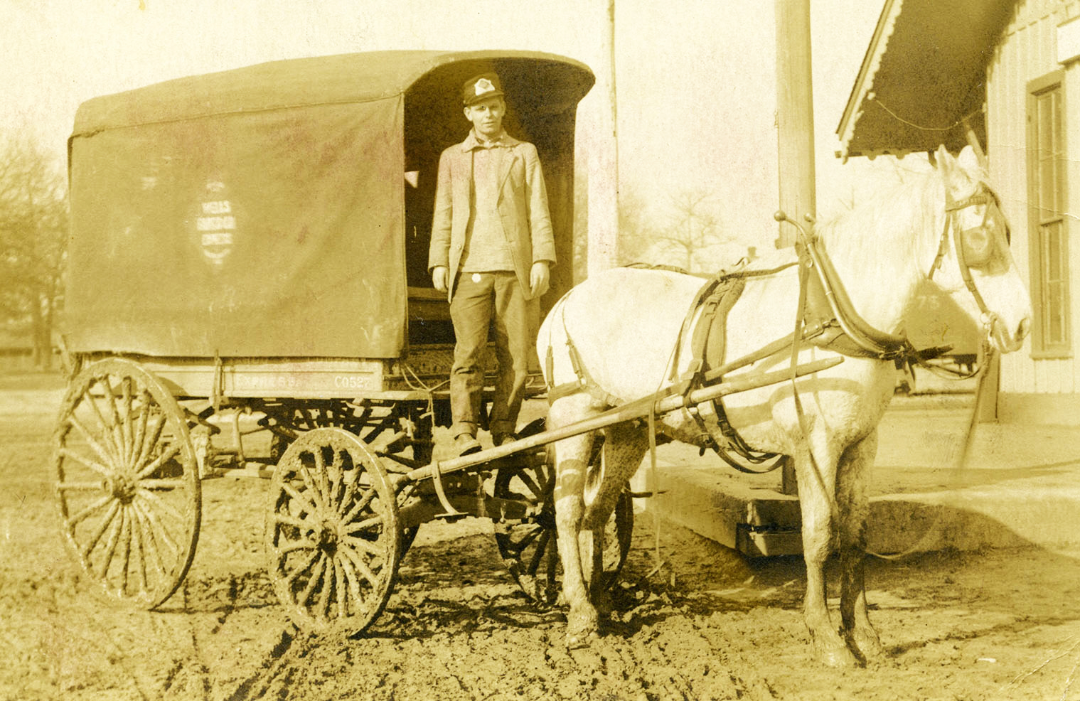 Wells Fargo express wagon with canvas cover pulled by single white horse. Man stands on driver's seat looking at camera. Rutted dirt road in foreground. Building background right. Image is black and white.