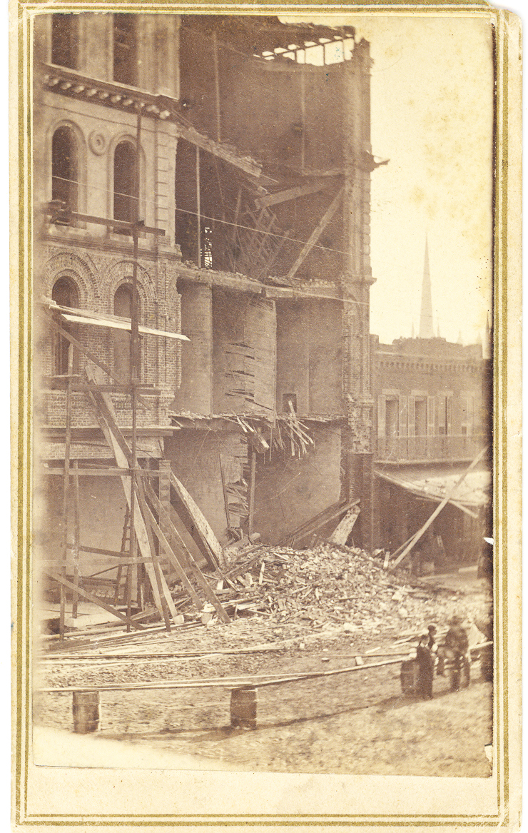 A three story brick building photographed with fallen front façade, and collapsed roof and floors. Piles of brick, stone and rubble surround building. Several men, backs to camera, survey the devastation. Image is black and white.