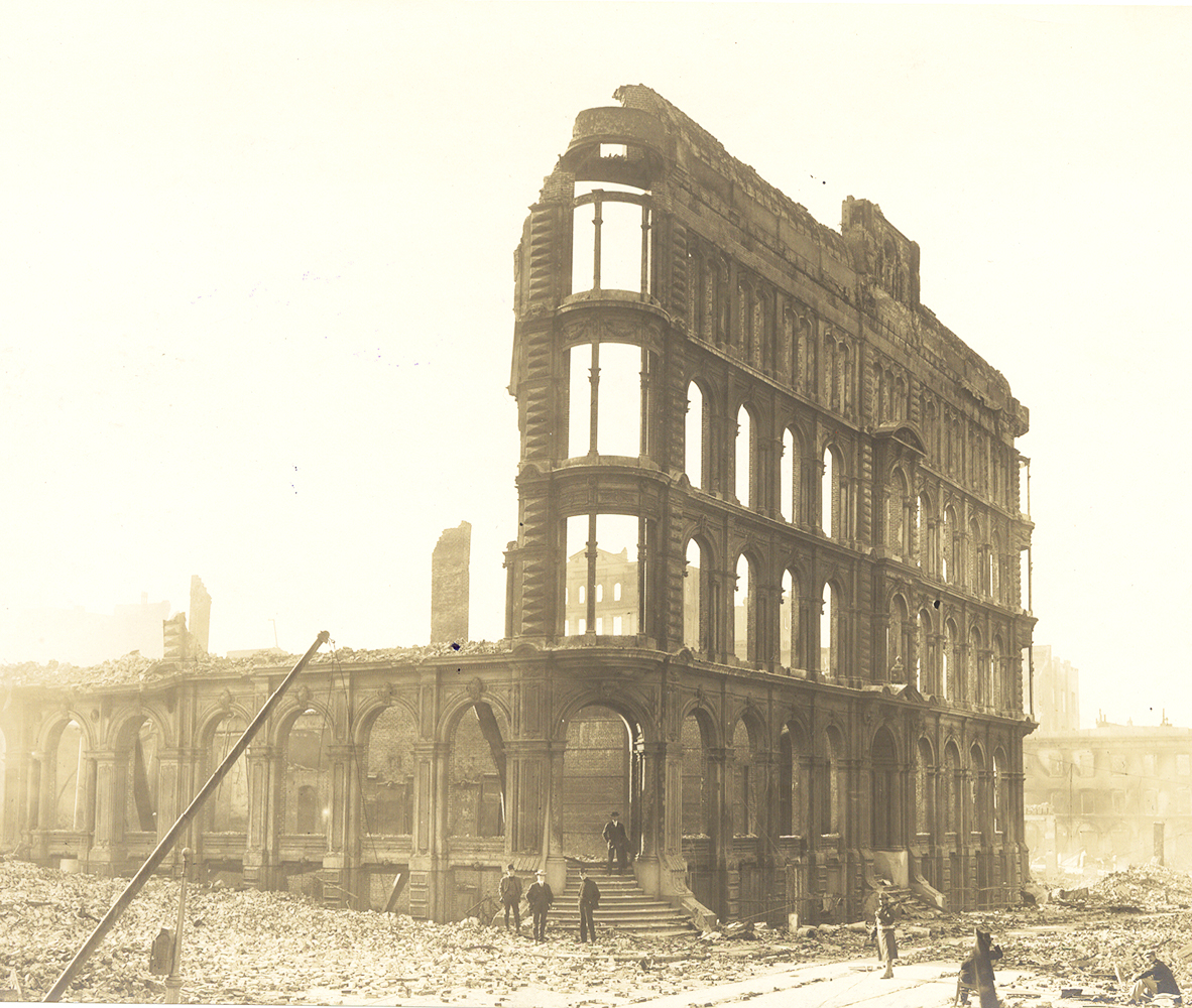 Burned out façade of four story building in ruins with piles of brick and debris in foreground. Several men stand near building. An armed soldier patrols the street. Smoke and haze appear in the air. Image is black and white.