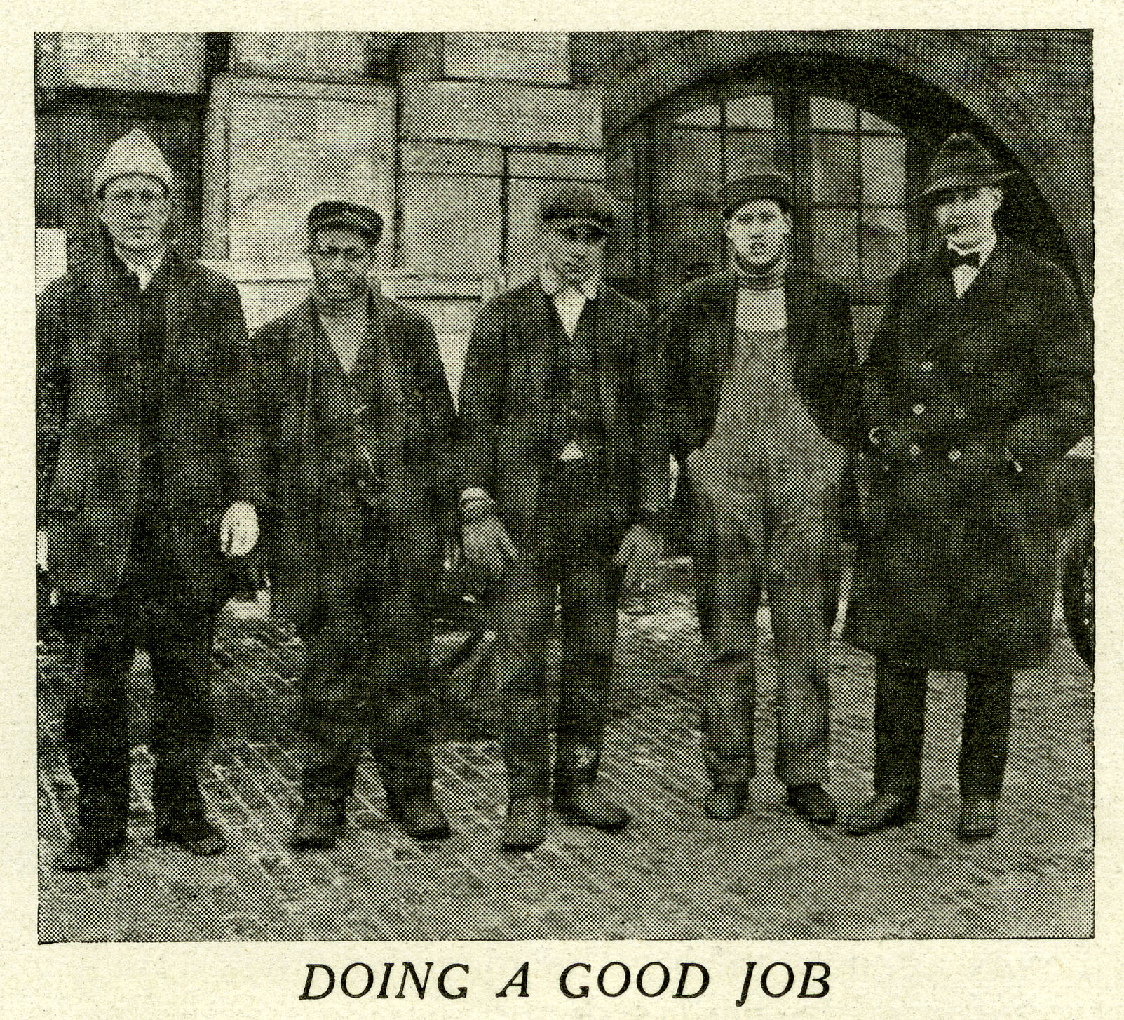 Group of five men standing on train platform. All are dressed in worker's clothes of overalls, pants, jackets and caps. Brick building and pile of wood boxes and crates behind them. Caption below reads Doing a good job. Image is black and white.