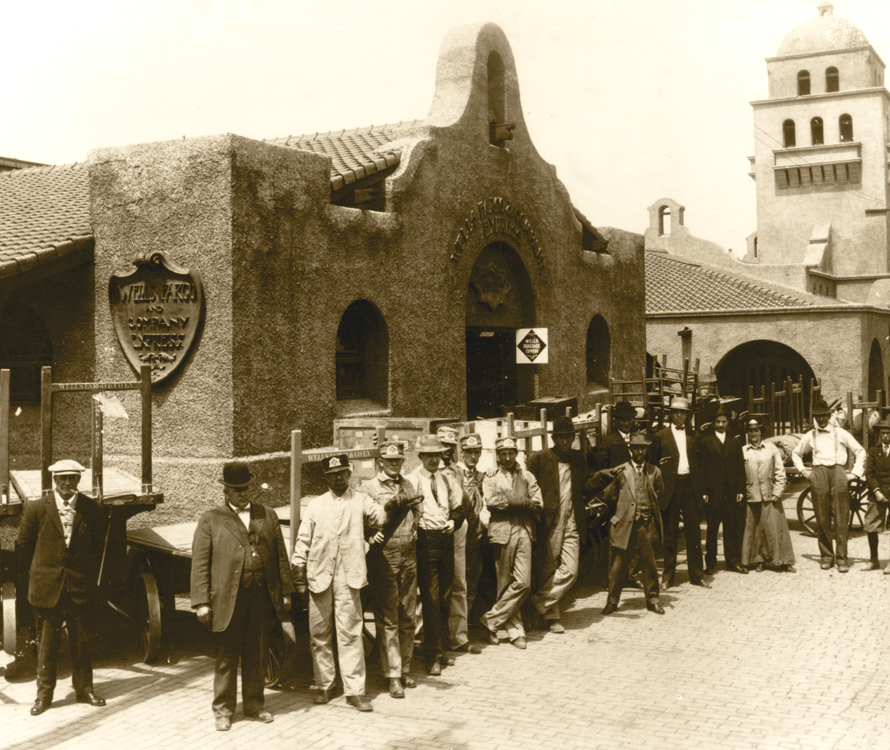 Fifteen men and one woman in overalls and other work clothes stand outside a spanish-style stucco railroad station building with sign reading Wells Fargo and Company Express. Wheeled carts crowd rail platform. Large tower of main station in background. Image is black and white.