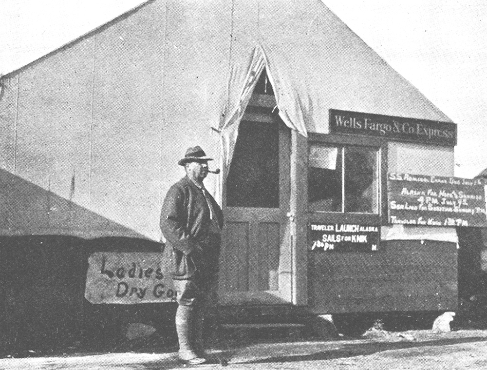 Man stands outside canvas tent with wooden door. Handwritten signs on tent include one reading Ladies Dry Goods. Wells Fargo & Co's Express sign hangs on canvas tent. Image in black and white.