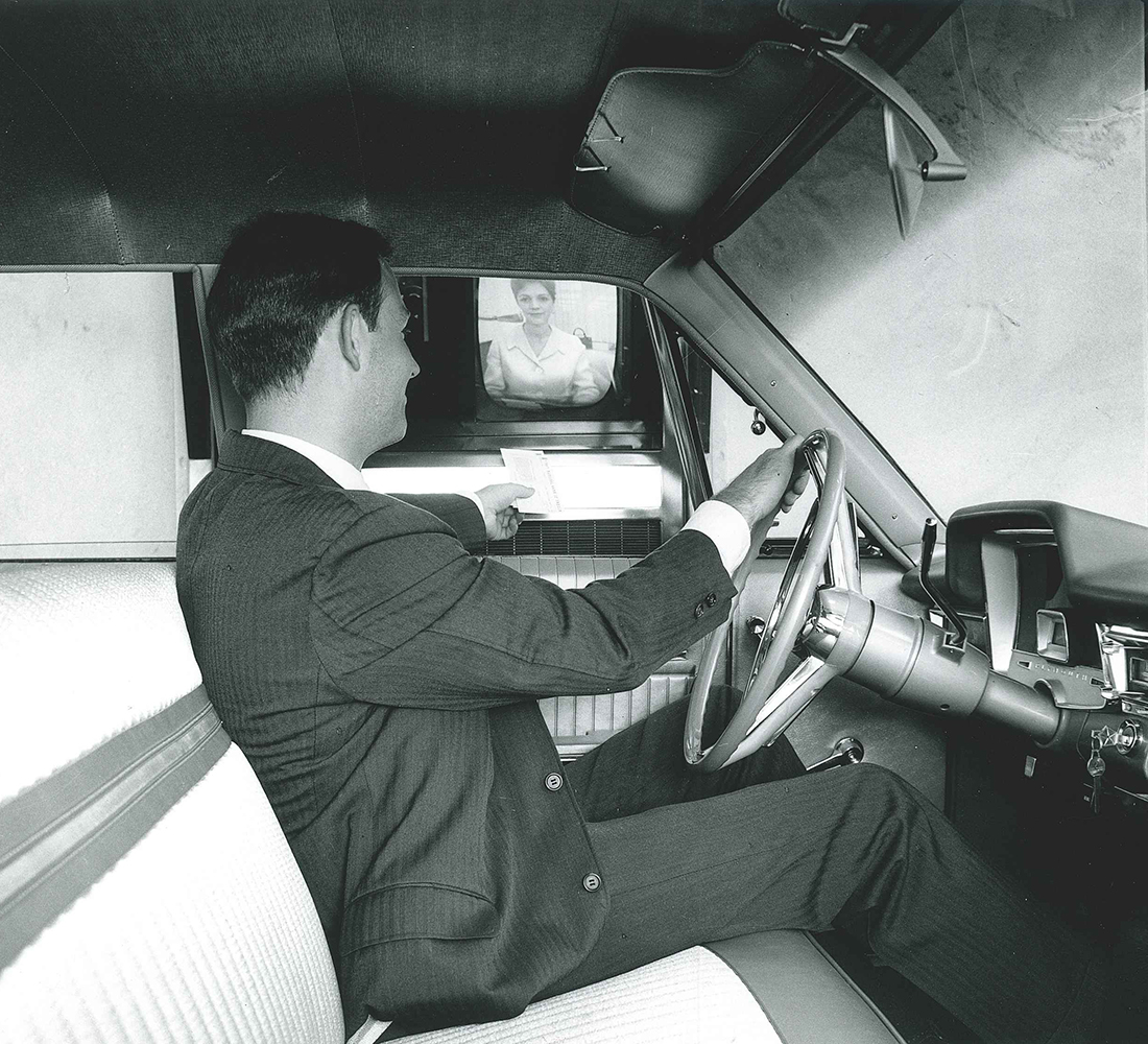 Man wearing suit sits in drivers seat of automobile. He holds a piece of paper at open driver's side window, looking out of the car to his left. His right hand is on the steering wheel. Bank teller is shown on small television screen to his left. Photo is taken through the passenger window of car. Image is black and white.