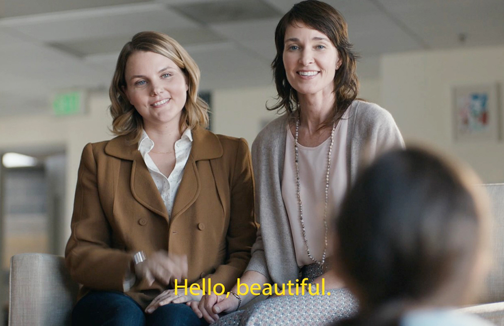 Advertising still photo of two smiling women with shoulder-length hair sign words in American Sign Language. They stand side by side and face the camera. Small girl with pony tail sits on floor in front of them, her back to the camera. Closed caption reads Hello beautiful.