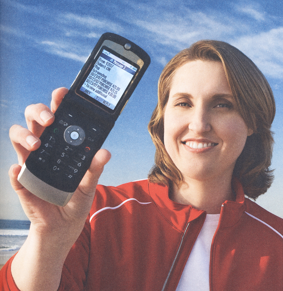 Smiling woman in red jacket holds up open flip phone to the camera. Text message shown includes some numbers representing banking transactions. Blue sky and beach form background. Image is color.