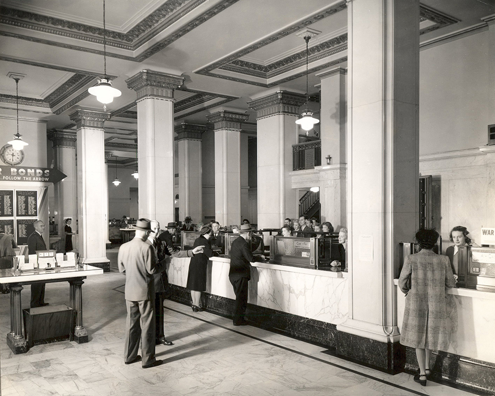 Bank branch interior shows multiple tellers behind teller counter, facing camera. Male and female customers face tellers, backs to camera. One man waits in line. All are dressed in hats, coats, suits and dresses. Banners on bank wall read War Bonds. Image is black and white.