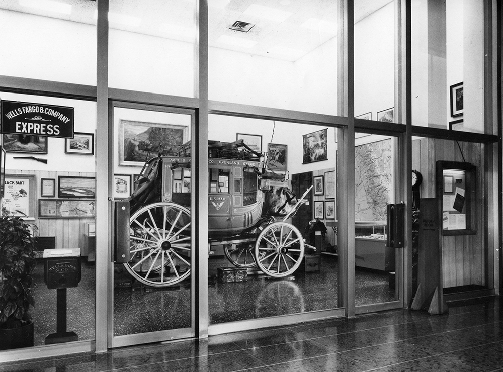 Room of historical artifacts including a stagecoach is contained behind a glass wall. Also visible surrounding stagecoach are framed images, maps, and signs. Image is black and white.