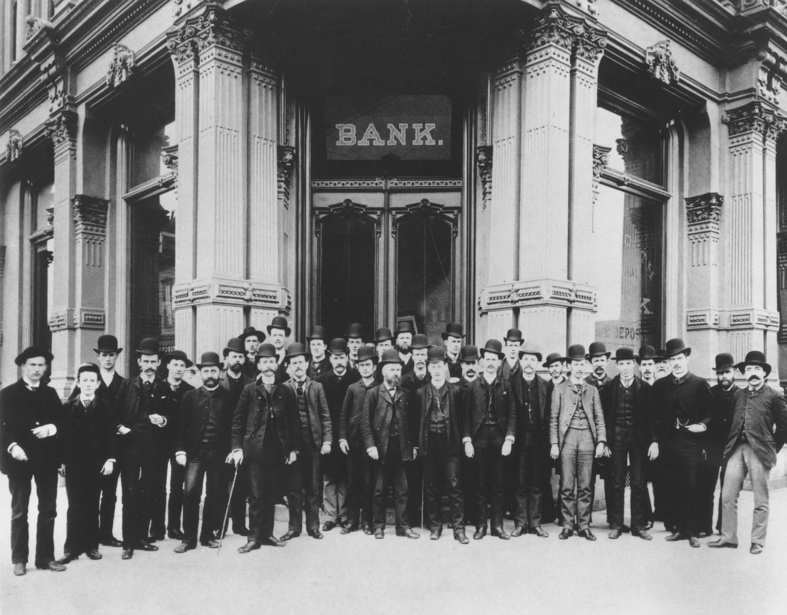 Group of thirty men in business attire suits pose in front of stone building on corner. The word Bank is visible above the front door. Image is black and white.