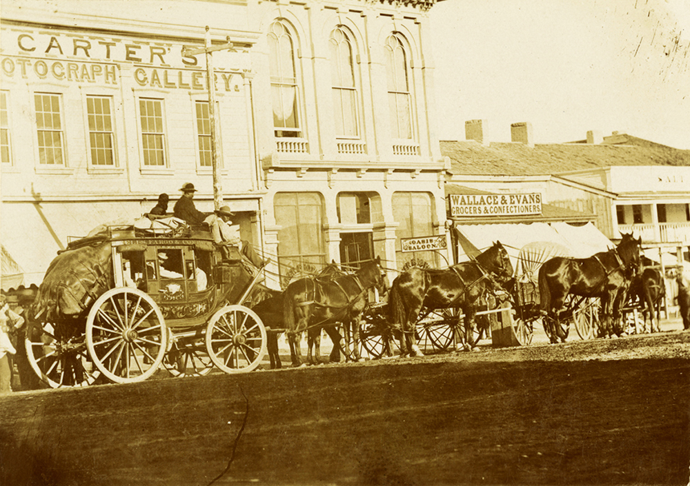 Streetview of stagecoach drawn by six horses shown facing left to right on unpaved city street. Driver and one additional person sit atop front of stage, directly above and behind horses. Several other passengers visible through stage windows. A number of stone commercial buildings in background, including one with signage reading Carters photograph gallery and another that reads Wallace & Evans Grocers & Confectioners. Image is black and white.