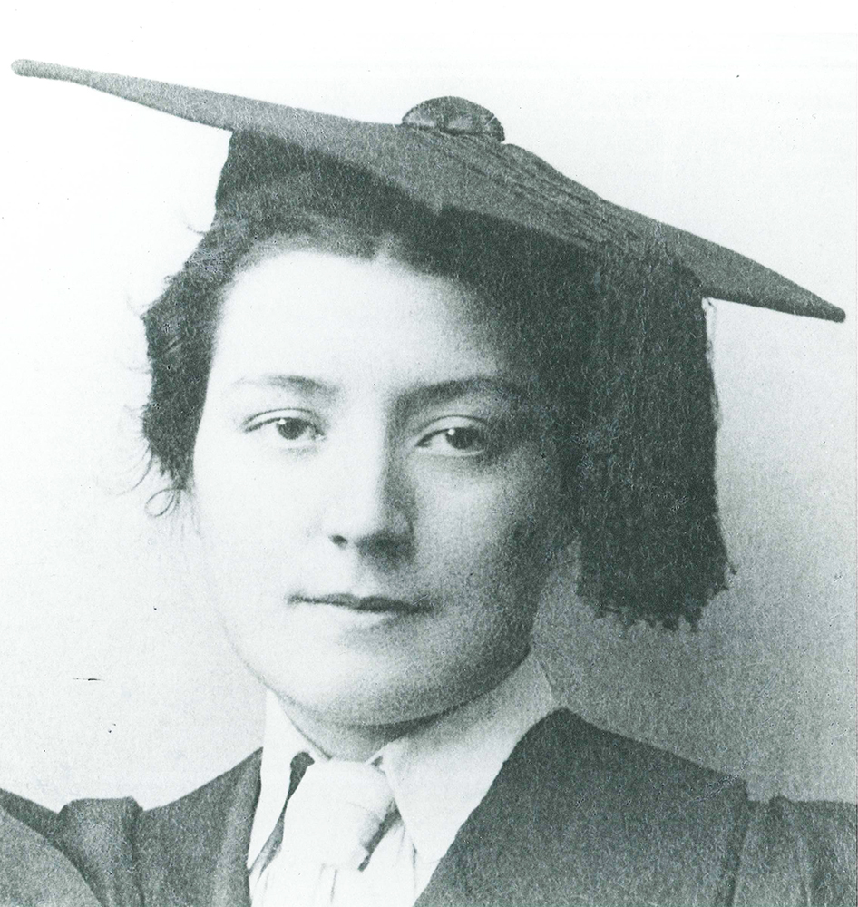 Portrait of dark-haired young woman in graduation cap and gown. She looks directly at the camera. Image is black and white.
