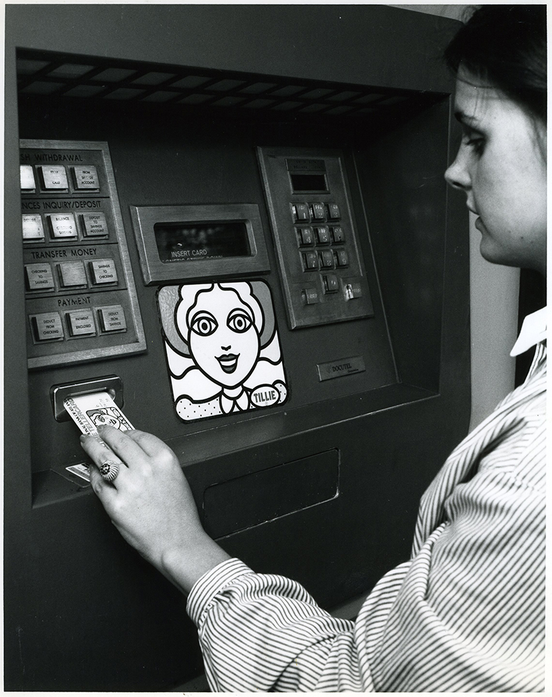A woman stands in front of bank ATM inserting her card with left hand. A cartoon character woman's face is shown on the machine. Character wears a name tag labeled Tillie. Image is black and white.