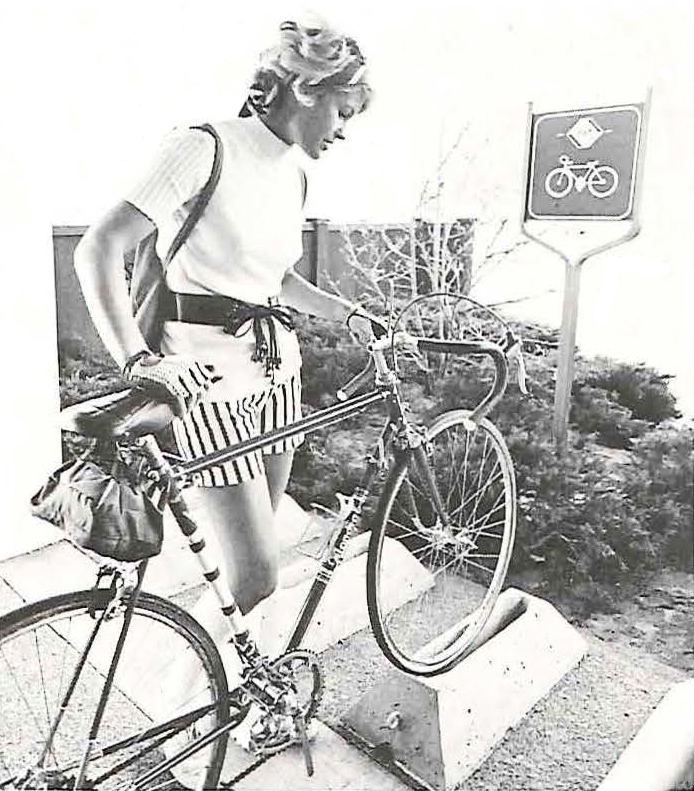 Young woman wearing shorts and summer clothing placing her bicycle's front wheel in a bike rack. Image is black and white.