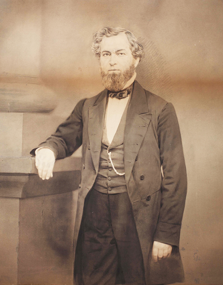 Portrait of bearded man with wavy hair standing while looking slightly right. He leans his right elbow and hand on a pillar. His left hand rests at his side. Dress is dark suit and tie, with prominent gold watch chain across his vest. Image is black and white.