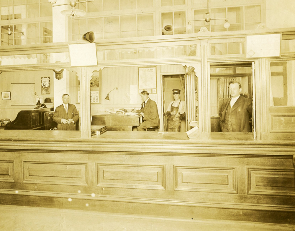 Photo shot inside building showing four men standing or sitting at work behind a tall wooden counter. Three are dressed in suits and ties, and one wears coveralls work clothing and hat. Several desks, calendars, and office equipment also behind the counter. Image is black and white.