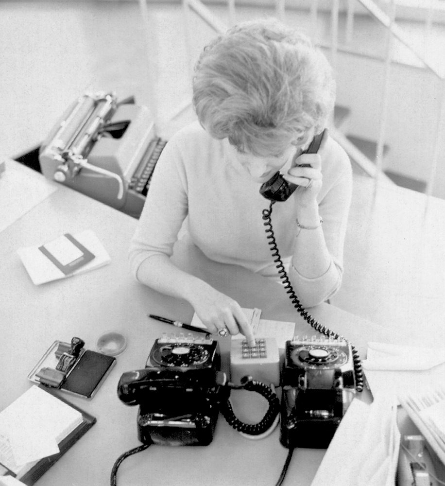 Overhead view of blonde woman sitting at desk holding phone receiver to her ear with left hand while keying in numbers on pushbutton keypad with her right hand. Other phone and office equipment sits nearby on her desk top. Image is black and white.