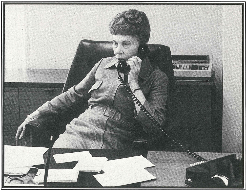 Woman with dark hair dressed in business attire leaning back in her chair at a desk while talking on the telephone. Various papers strewn on desk in front of her. Image is black and white.
