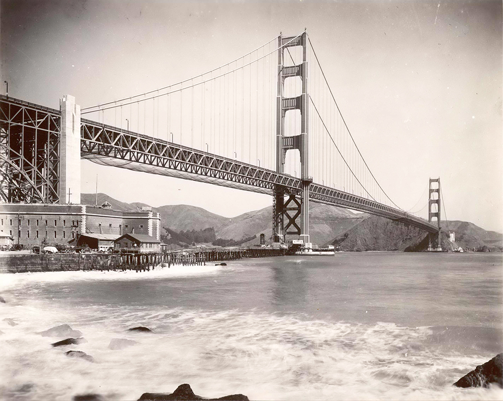Photo looking up at Golden Gate Bridge from water level near south anchorage. Surf and shoreline in foreground. Image is black and white.