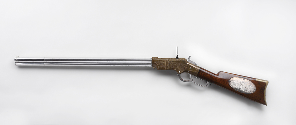 Rifle shown in profile, pointing right. Has silver plated barrel and dark polished wood stock. There is an oval shaped engraved silver plaque on stock. Image is color.