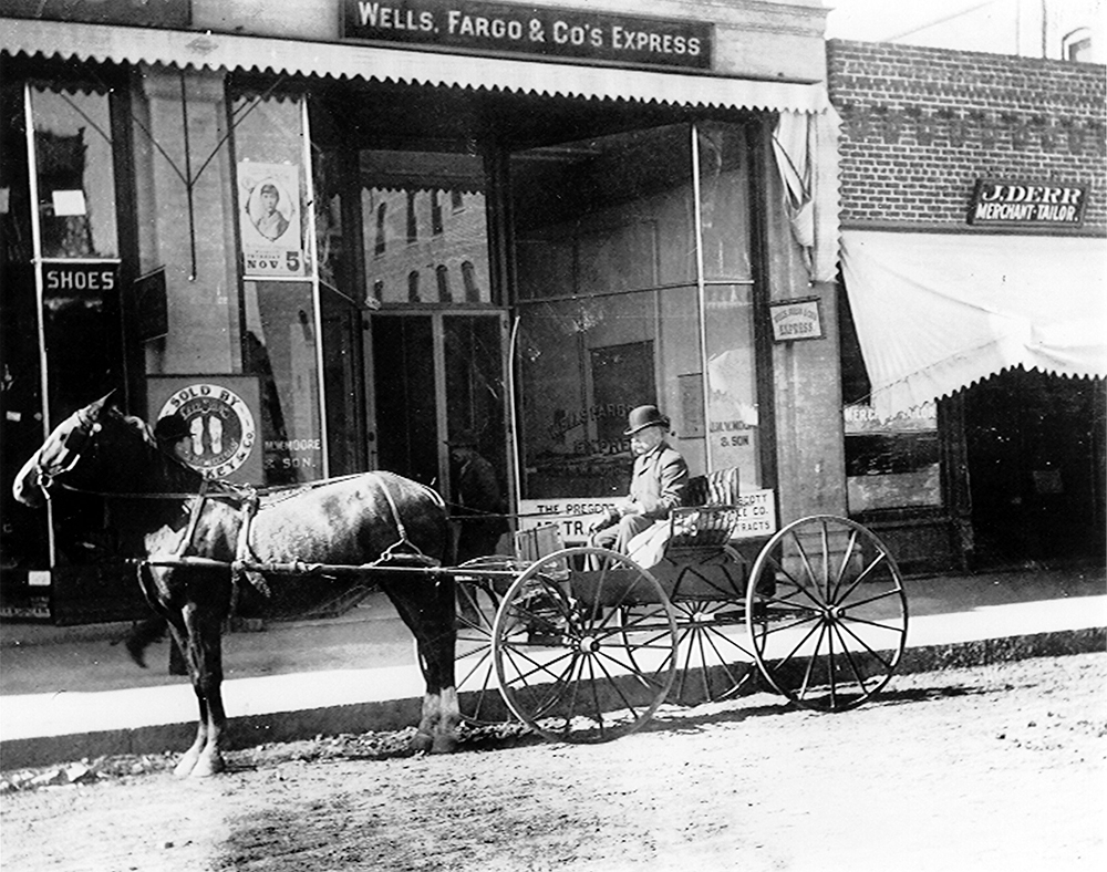 A street scene with a carriage drawn by a single horse parked at the curb. The driver is wearing a black suit jacket and boller hat. A sign above the glass windows of the store front reads: Wells Fargo & Co.'s Express. Image in black and white.