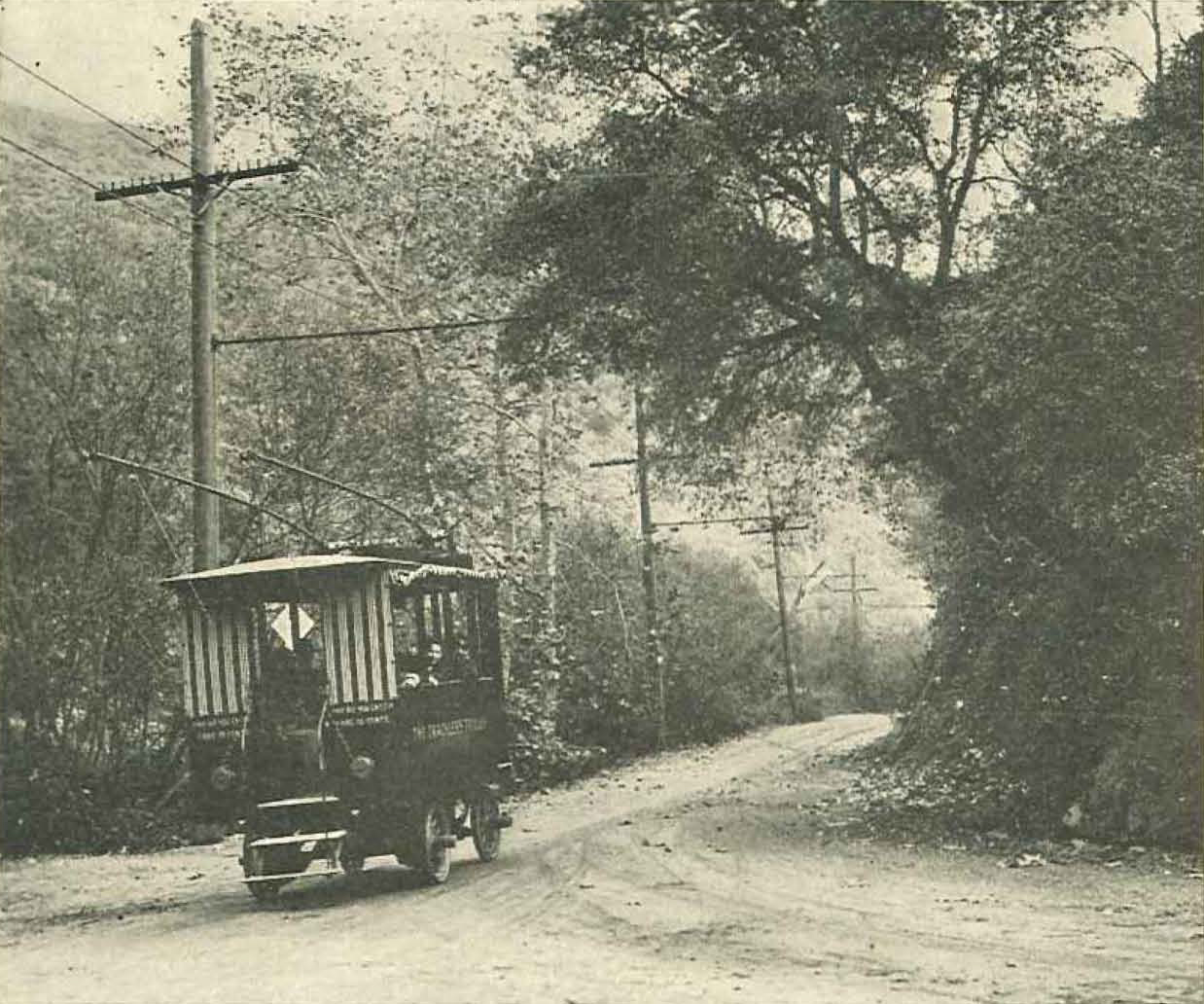 A small trolley descends on a curvy dirt road. Metal trolley has open sides of striped rolled canvas. Electrical poles extend from roof, reaching wires strung overhead. Two women lean out left side of trolley. Image is black and white.
