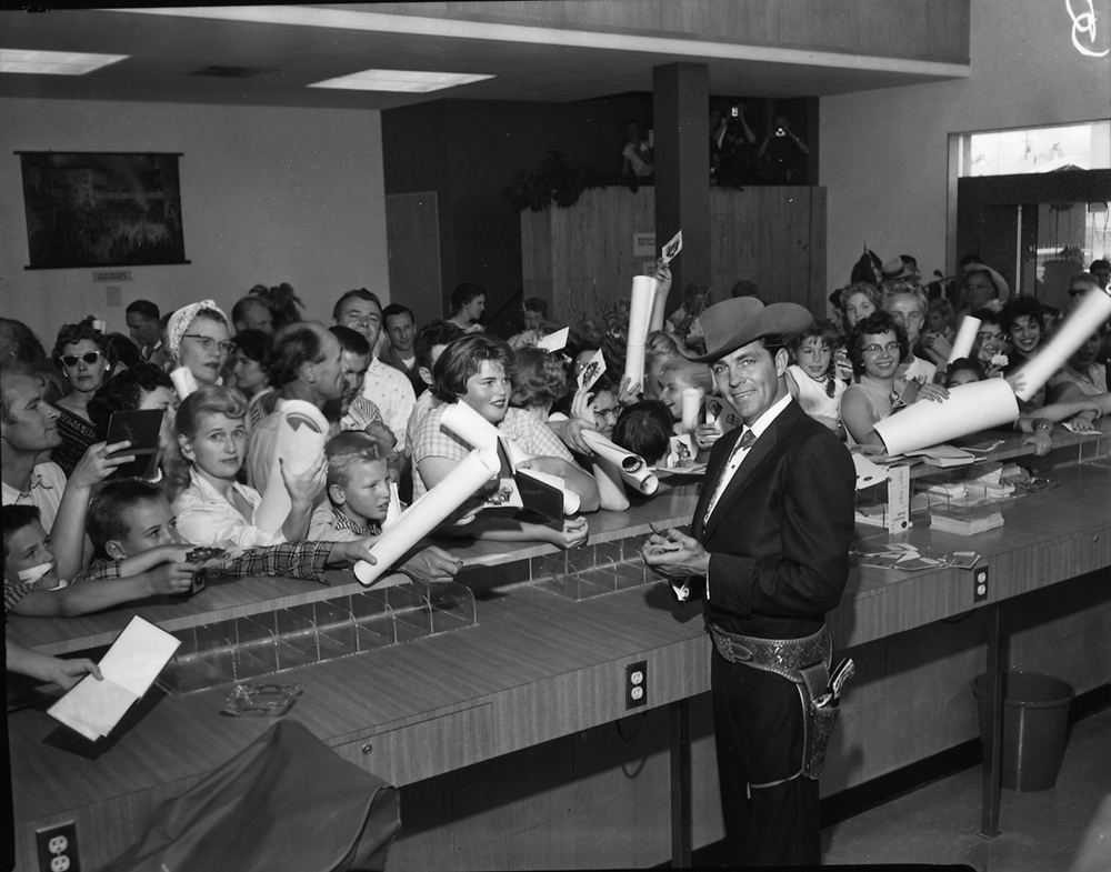 Photo of television celebrity Dale Robertson dressed in western attire with hat and holstered gun. He turns to the camera while signing autographs for crowd of fans on opposite side of bank counter inside a Wells Fargo branch. Image is black and white.