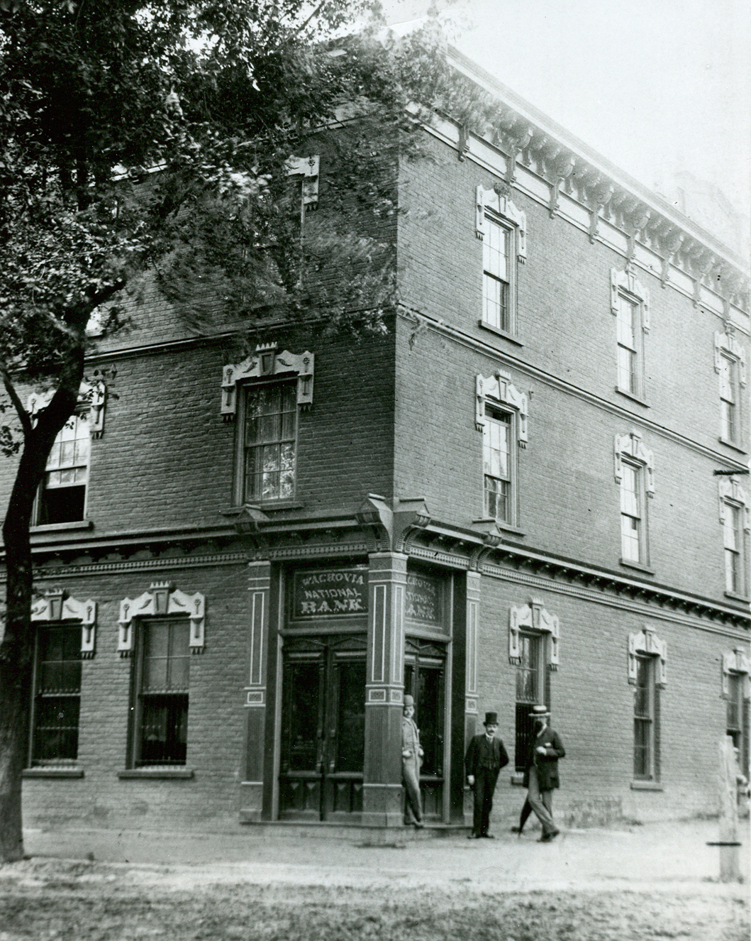 Three story brick building on corner with words Wachovia National Bank over door. Three men in business attire stand just outside door. Large tree foreground left. Image is black and white.