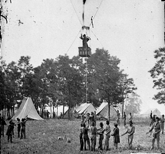 Union Army officer ascends in observation balloon's wicker basket. Six soldiers hold ropes tethering the balloon to the ground. Army tents and trees in background. Image is black and white.