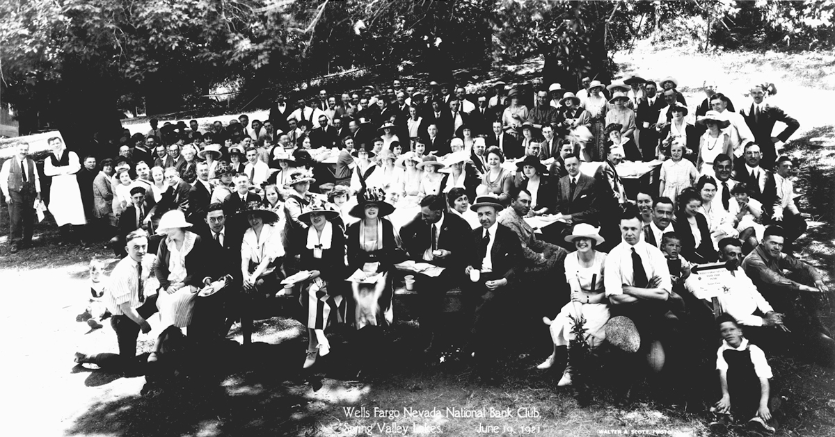Panoramic photo of large group of several hundred men, women, and children sit or stand at long tables set for picnic meal outdoors in wooded area. Label reads Wells Fargo Nevada National Bank Club Spring Valley Lakes June 19, 1921 at bottom of photo. Image is black and white.