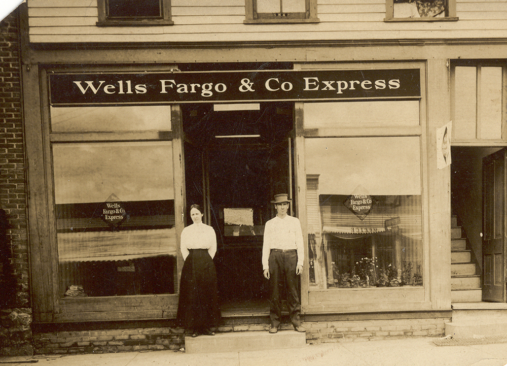 Photo of man and woman standing on either side of door in front of small commercial building. Each wears white shirt and dark skirt or pants. Large sign saying Wells Fargo & Co. Express over door, with diamond-shaped Wells Fargo & Co. express signs painted on windows either side of door.