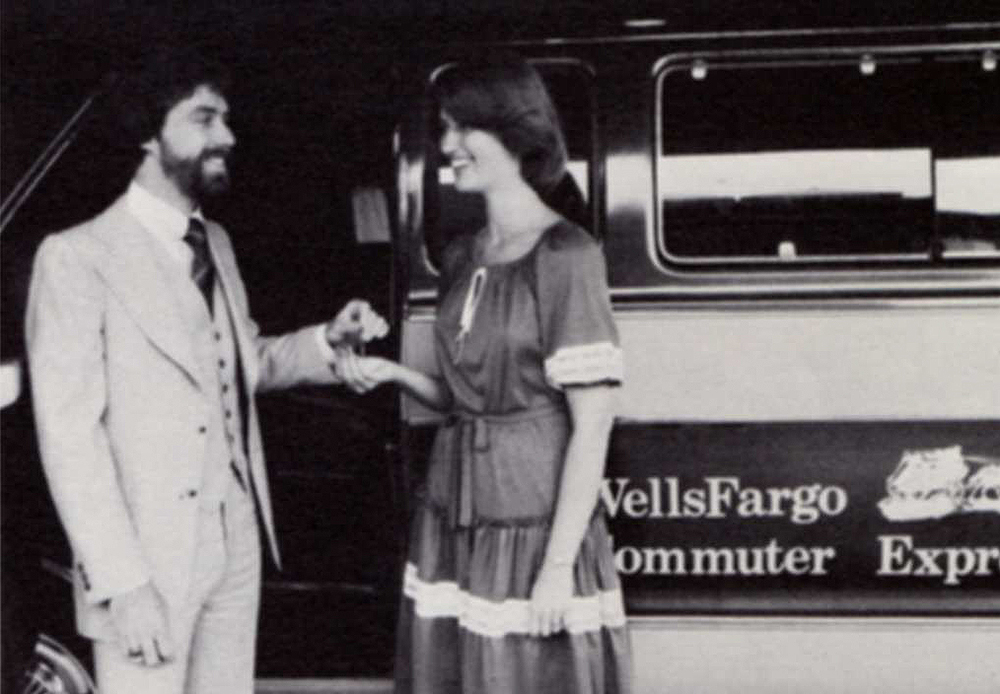 Bearded Caucasian man in business suit and Caucasian woman in dark dress stand in front of passenger van with sign reading Wells Fargo commuter express. Image is black and white.